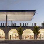 Stazione moderna con treno, passanti sul ponte e negozi illuminati sotto gli archi al tramonto.