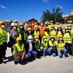 Gruppo di persone con caschi gialli e giubbotti catarifrangenti in un cantiere sotto un cielo blu.