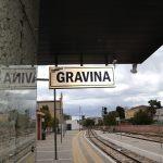 Cartello della stazione ferroviaria di Gravina, binari deserti sotto cielo nuvoloso.
