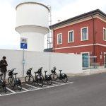 Parcheggio bici condivise vicino a edificio rosso e serbatoio bianco, uomo con telefono, cielo nuvoloso.