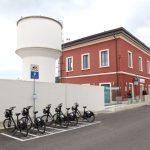 Parcheggio biciclette bike sharing, edificio rosso e serbatoio idrico bianco sullo sfondo, giornata nuvolosa.