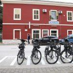 Biciclette nere parcheggiate davanti alla stazione di Altamura, edificio rosso e auto sullo sfondo.
