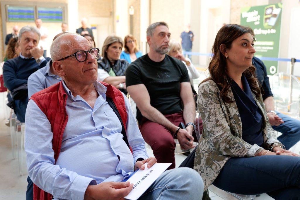 Pubblico attento durante un evento, con diverse persone sedute in ascolto in un ambiente luminoso.