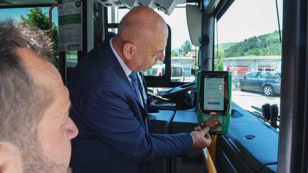 Un uomo in giacca e cravatta utilizza un lettore di carte sul bus, mostrando un pagamento elettronico per il trasporto.